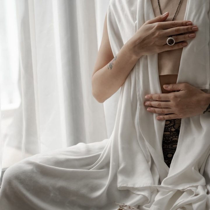 Eine Person in weißem Stoff sitzt und berührt sanft ihren Bauch.